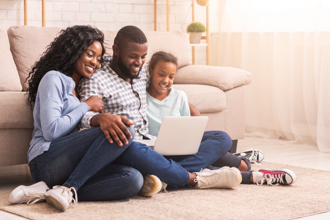 Planning vacation together. African father, mother and daughter using laptop at home, sitting on floor, copy space