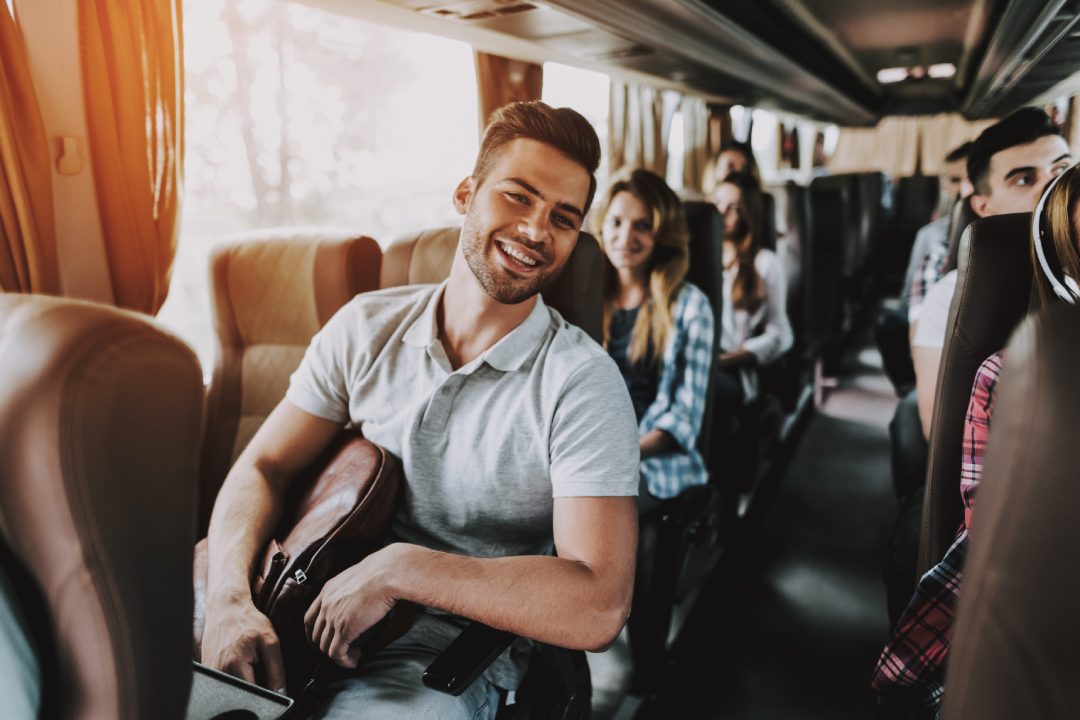 Young Handsome Man Relaxing in Seat of Tour Bus. Attractive Smiling Man Sitting on Passenger Seat of Tourist Bus and Holding Backpack. Traveling and Tourism Concept. Happy Travelers on Trip