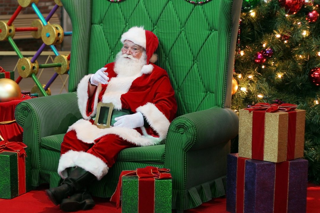 authentic santa claus sitting in large green chair surrounded by presents and decorations