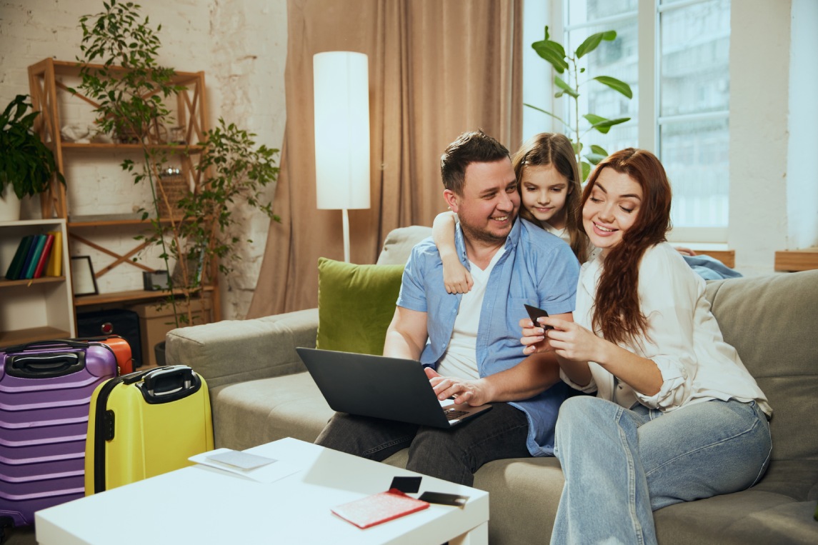 Young family, mother, father and daughter sitting at home with suitcases, planning trip, making online hotel reservation, booking tickets. Concept of vacation, traveling, booking services