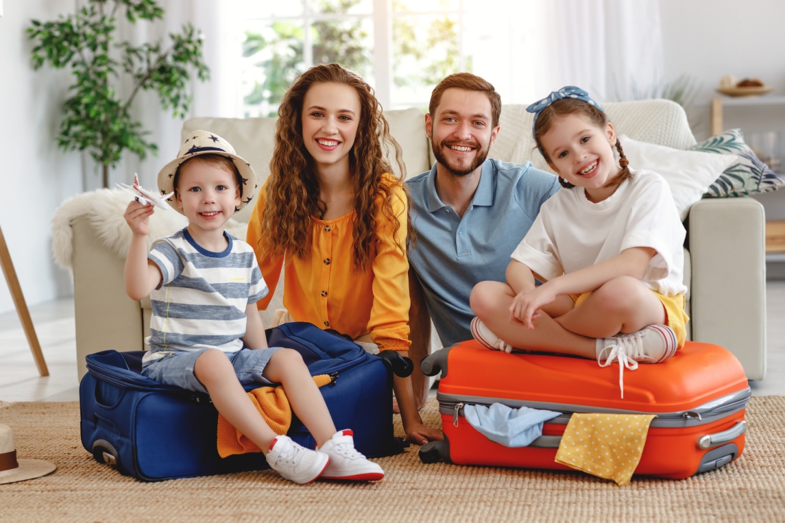 Happy parents with children laughing at camera while sitting on floor near sofa with suitcases near at home